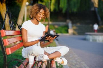 Fototapeta premium Young curly black smiling female with a digital pad on the bench in the park; charming cheerful Brazilian girl with Afro hair using tablet pc while sitting on the wooden bench on a summer day