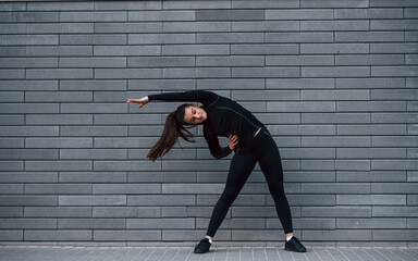 Young sportive girl in black sportswear outdoors doing stretching near gray wall
