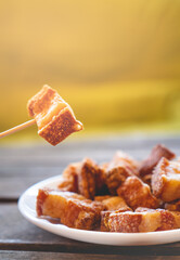 A toothpick holding a piece of fried pork rinds called “Torreznos” over a small white round fried pork rinds plate on a dark wooden table. Photo with selective focus. 