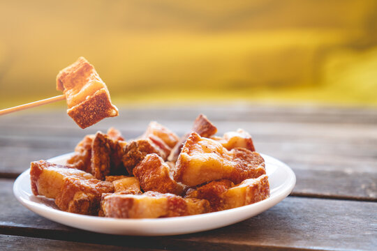 A toothpick holding a piece of fried pork rinds called &ldquo;Torreznos&rdquo; over a small white round fried pork rinds plate on a dark wooden table. Photo with selective focus. 