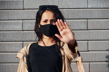 Showing stop gesture by hand. In protective mask. Beautiful brunette with curly hair and in black clothes outdoors near wall