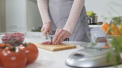 Obraz na płótnie Canvas Woman cutting raw meat on small pieces with kitchen knife cutting board. Woman puts pieces of raw meat in a glass bowl. Female cutting red beef filet on wooden board, holding knife in hand.
