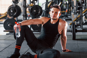Fototapeta na wymiar Taking a break and drinking water. Young sportive strong man in black wear have workout day in gym