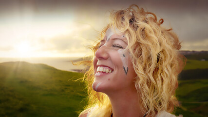 Beautiful, carefree smiling girl with blonde wavy hear and Celtic symbols painted on her face enjoying the sunset at the coast at dusk.