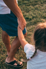 Father and daughter hold hands. The child's hand and the man's hand hold each other. Dad and daughter walk together holding hands on the grass in the summer at sunset