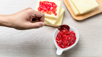 Strawberry Jam in white ceramic cup placed on wooden table with blurred bread background. Food close up