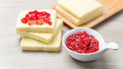 Strawberry Jam in white ceramic cup placed on wooden table with blurred bread background. Food close up