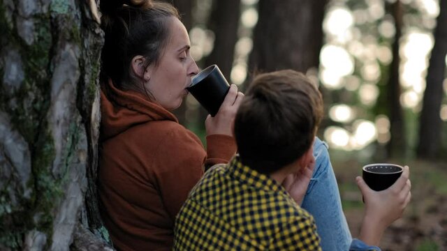Back view of Mother with son relaxing in the forest and drinking hot herbal tea from a thermos. Stay cation concept

