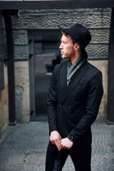 Elegant young man in black hat and clothes with umbrella in hands outdoors in the city