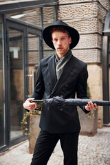 Obraz premium Elegant young man in black hat and clothes with umbrella in hands outdoors in the city