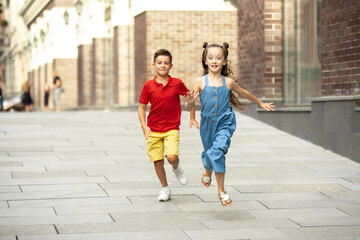 Two smiling kids, boy and girl running together in town, city in summer day. Concept of childhood, happiness, sincere emotions, carefree lifestyle. Little caucasian models in bright clothes.