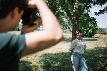 Girl who is posing near tree in nature having a photo session. Blurred photographer taking photos with professional camera of female model in forest.