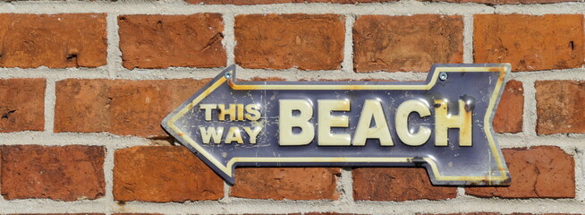This way beach. Old rusty metal sign on a red brick wall. Blue with white lettering. 
Path to the beach, signpost and  tin sign with an arrow pointing to the left.

