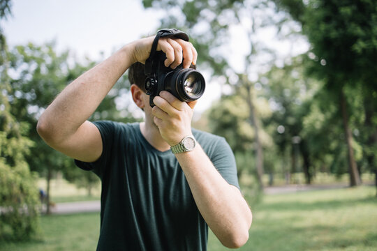 Front view of man taking photos in nature. Portrait of photographer looking through camera viewfinder and taking pictures while standing in park.