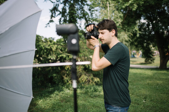Photographer taking pictures in park using professional camera. Man looking through camera viewfinder and taking pictures while standing next to flash on stand outdoor.