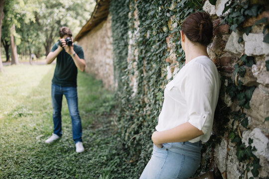 Blurred man with professional camera taking pictures of girl. Out of focus photographer shooting woman who is leaning against wall with ivy.
