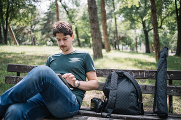 Obraz premium Man sitting on bench in park and using tablet. Young male photographer resting on wooden seat with backpack in park while looking at tablet.