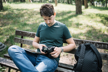 Naklejka na ściany i meble Man sitting on bench in park and looking at his camera. Handsome photographer sitting on seat in park with backpack and using DSLR camera.