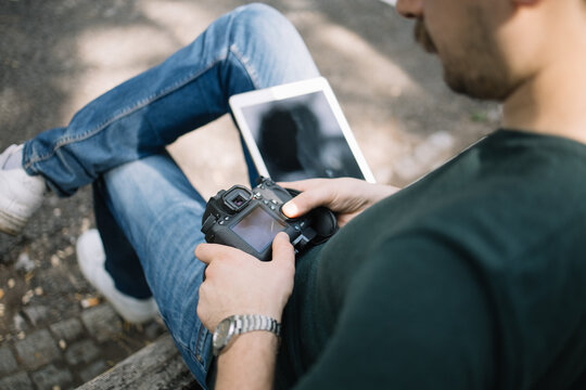 Man sitting on bench with tablet and professional camera. High angle view of photographer holding camera and having tablet in his lap while sitting on seat outdoor.