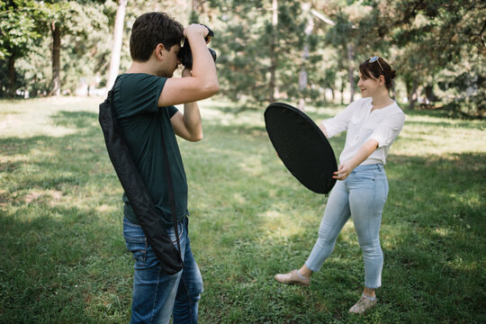 Side view of photographer taking pictures of woman. Man with professional camera taking photos of woman who is holding reflector in nature.