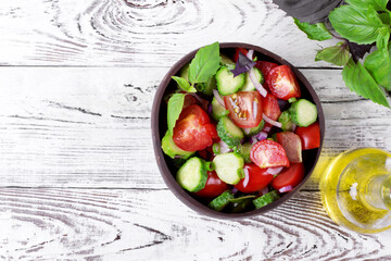 Salad with cucumbers, tomatoes, basil, red onion and olive oil in a wooden bowl on the white table