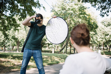 Photographer taking photos of out of focus female model. Man using professional camera and reflector photoshooting back of blurred girl in park.