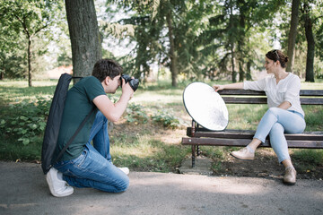 Side view of girl sitting on bench while photographer is taking photos of her. Man with professional camera shooting female model with reflector in park.