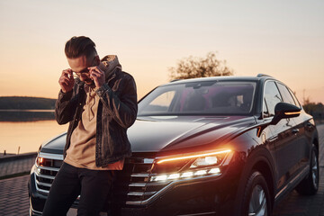 Handsome unshaved man in fashionable clothesleaning on his his black car