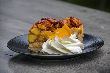 Black plate with apple pie and whipped cream with a slic of orange on a glassy wooden table