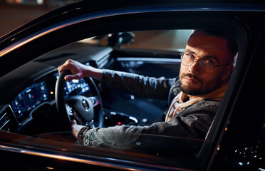 Side view of handsome unshaved man in fashionable clothes that riding his automobile