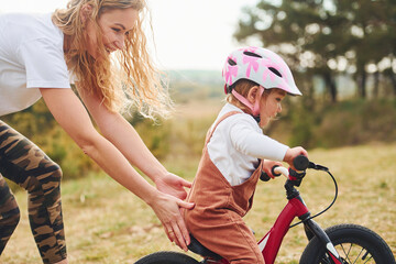 Naklejka premium Mother in white shirt teaching daughter how to ride bicycle outdoors
