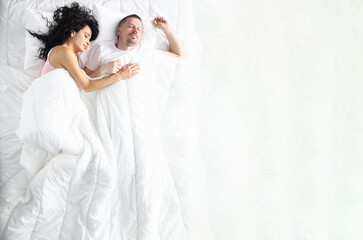 Husband and wife sleep soundly under a white blanket. A man sleeps on his back, and a woman on his...