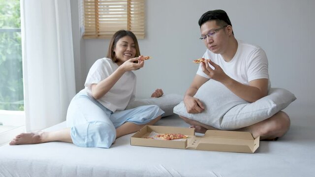 Normal shot : Young attractive happy asian married couple eat pizza, take-out food or fast food on bed feeling positive and relax on weekend quarantine lifestyle activity.