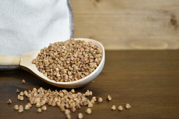 Buckwheat in a wooden spoon, next to a canvas sac with grain on a brown wooden background. Healthy food concept, vegetarian food. Copy space. Selective focus