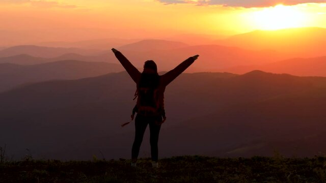 Young inspired woman raises her hands up standing on the top of a mountain against beautiful sunrise sky. Watching the sunset with beautiful landscape