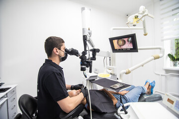 Delightful girl with patient bib on a dental chair and a dentist who sits next to her looks on her teeth using a dental microscope and holds a dental bur and a mirror.