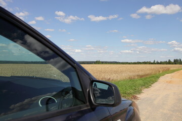 Fototapeta na wymiar Car side door glass and mirror on Golden field, blue sky with white clouds and rural sandy road beautiful background, eco car tourism in Europe countryside 