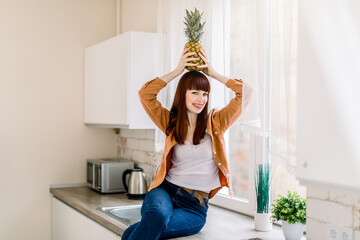 Shot of beautiful young woman in jeans and casual shirt, holding fresh pineapple on head and smiling to camera, posing at home kitchen sitting on the countertop. Healthy food, home interior
