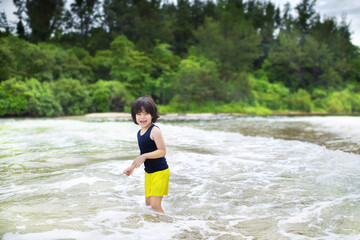 Cute Asian boy standing in the waves by the sea.Child having fun outdoor.Wildschooling young male playing in the water at the shore.
