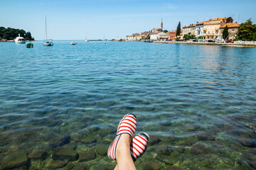 Woman's feet with espadrilles hanging over the transparent Adriatic sea water with croatian Rovinj city and boats on the background