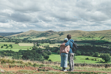 Fototapeta na wymiar Father is hugging his daughter and they areobserving beautiful field view on Edale village and Mam Tor at Peak District National Park, England, UK while hiking. Staycation concept of traveling local