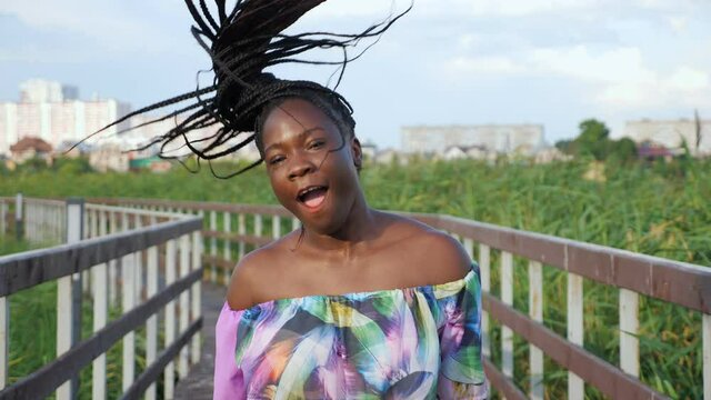 pretty young African-American lady in bright blouse shakes long dark hair with cornrows on wooden bridge among green reeds against city slow motion