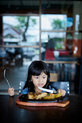 Little girl holding knife and fork cutting grilled Chicken steak on stoned plate in restaurant