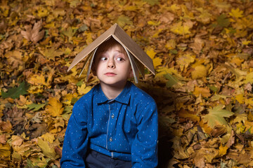 Scared boy sitting on fallen leaves in park and holding book over his head