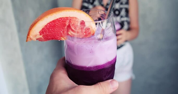 POV close up shot of a hand holding a fruity summer sweet violet purple alco cocktail with a metal straw and orange slice. Celebration and clinking with reflective transparent glass with girl in slow 