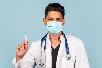 Cute  Indian / Asian doctor with a stethoscope and a syringe in his hands, in a white coat on a blue background.