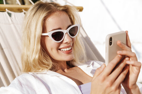 Image of charming smiling woman using cellphone while lying in hammock