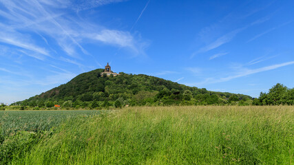Fototapeta na wymiar Green farm field at the foot of the mountains in sunny summer day. At the top is a monument to Emperor William. Porta Westfalica, Kaiser Wilhelm Memorial
