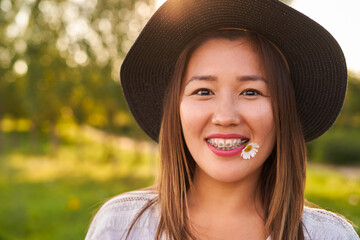 Braces on the teeth, and in the mouth a chamomile flower, a girl of Asian appearance in a white T-shirt and a dark hat, looks at the camera and smiles, blurred background