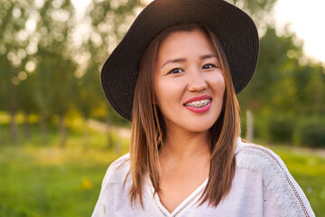 Braces on the teeth, a girl of Asian appearance in a white T-shirt and a dark hat, looking at the camera and smiling, blurred background